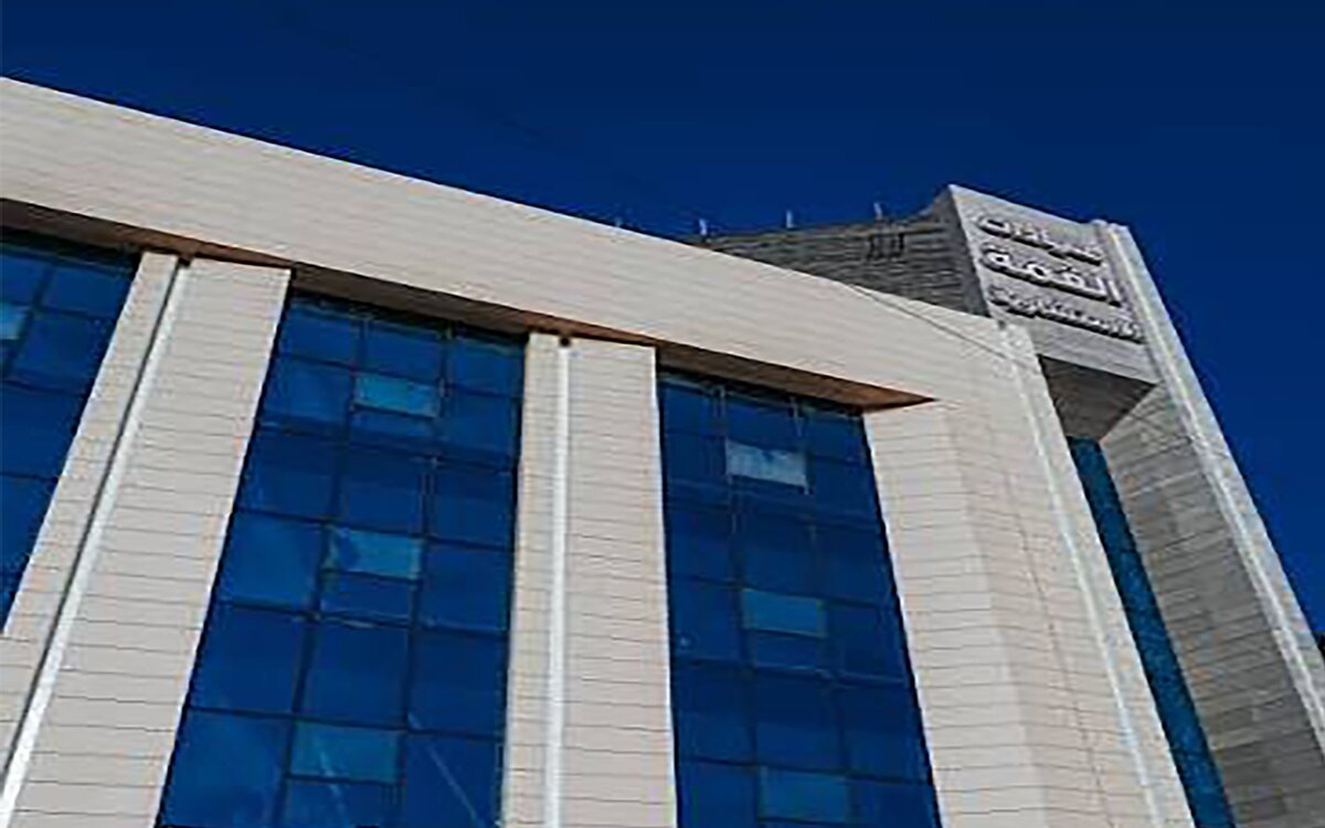 Exterior of the Al-Qimma Hospital building — a multi-storey stone facade with blue glazed window bays and Arabic signage on the corner tower