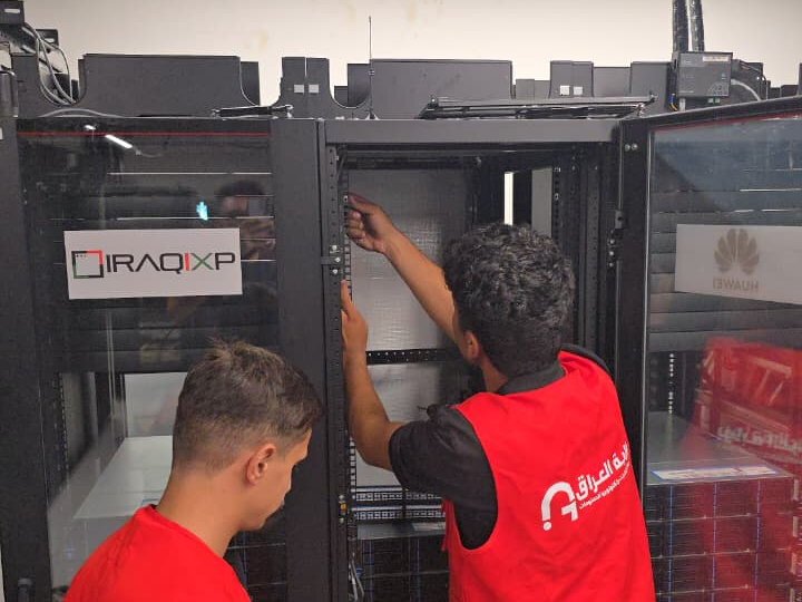 Two Iraq Gate technicians in branded vests working at an open server rack, beside a rack bearing an IRAQIXP label